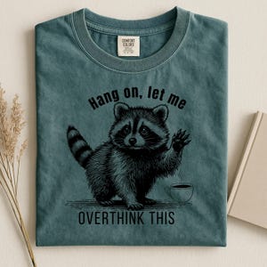 A dark teal t-shirt with a black graphic of a raccoon holding up a paw next to a steaming coffee cup, with the text "Hang on, let me OVERTHINK THIS."