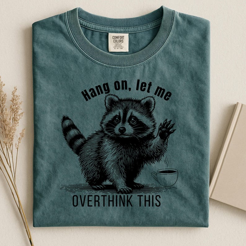 A dark teal t-shirt with a black graphic of a raccoon holding up a paw next to a steaming coffee cup, with the text "Hang on, let me OVERTHINK THIS."