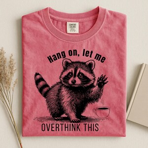 A bright pink t-shirt with a black graphic of a raccoon holding up a paw next to a steaming coffee cup, with the text "Hang on, let me OVERTHINK THIS."