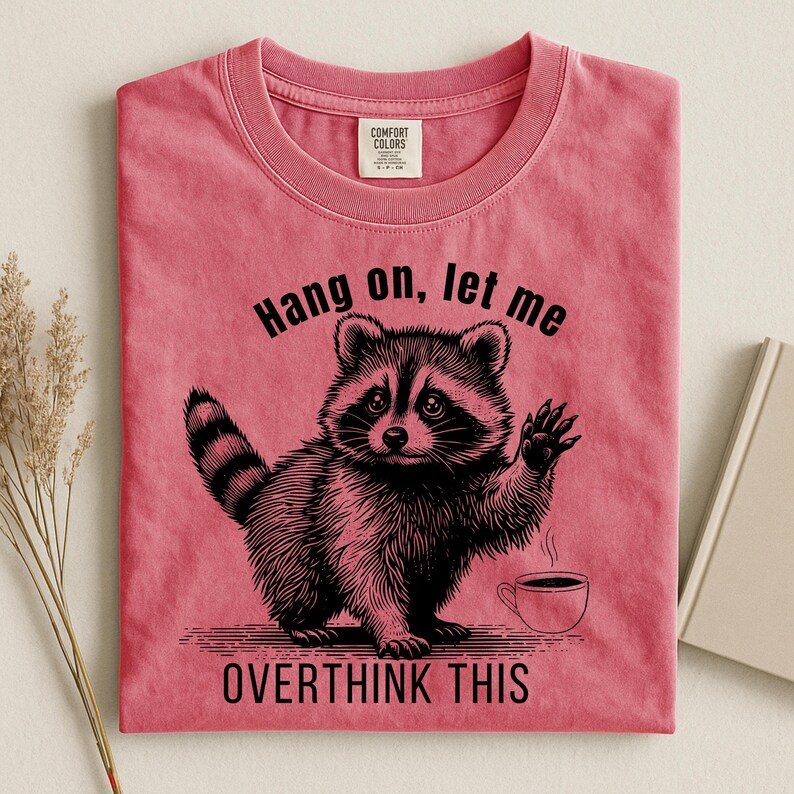 A bright pink t-shirt with a black graphic of a raccoon holding up a paw next to a steaming coffee cup, with the text "Hang on, let me OVERTHINK THIS."