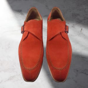 Könnte beinhalten: Ein Paar leuchtend orangefarbene Wildlederschuhe mit einem Monk-Strap-Schnallenverschluss. Die Schuhe haben eine runde Spitze und dekorative Nähte. Präsentiert auf einer Marmoroberfläche.