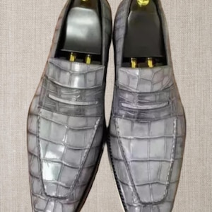 Op de afbeelding: Een paar grijze leren loafers met een krokodillenleertextuur. De schoenen hebben een klassiek ontwerp met een penny loafer-bandje en een ronde neus. Schoenbomen zijn ingevoegd.