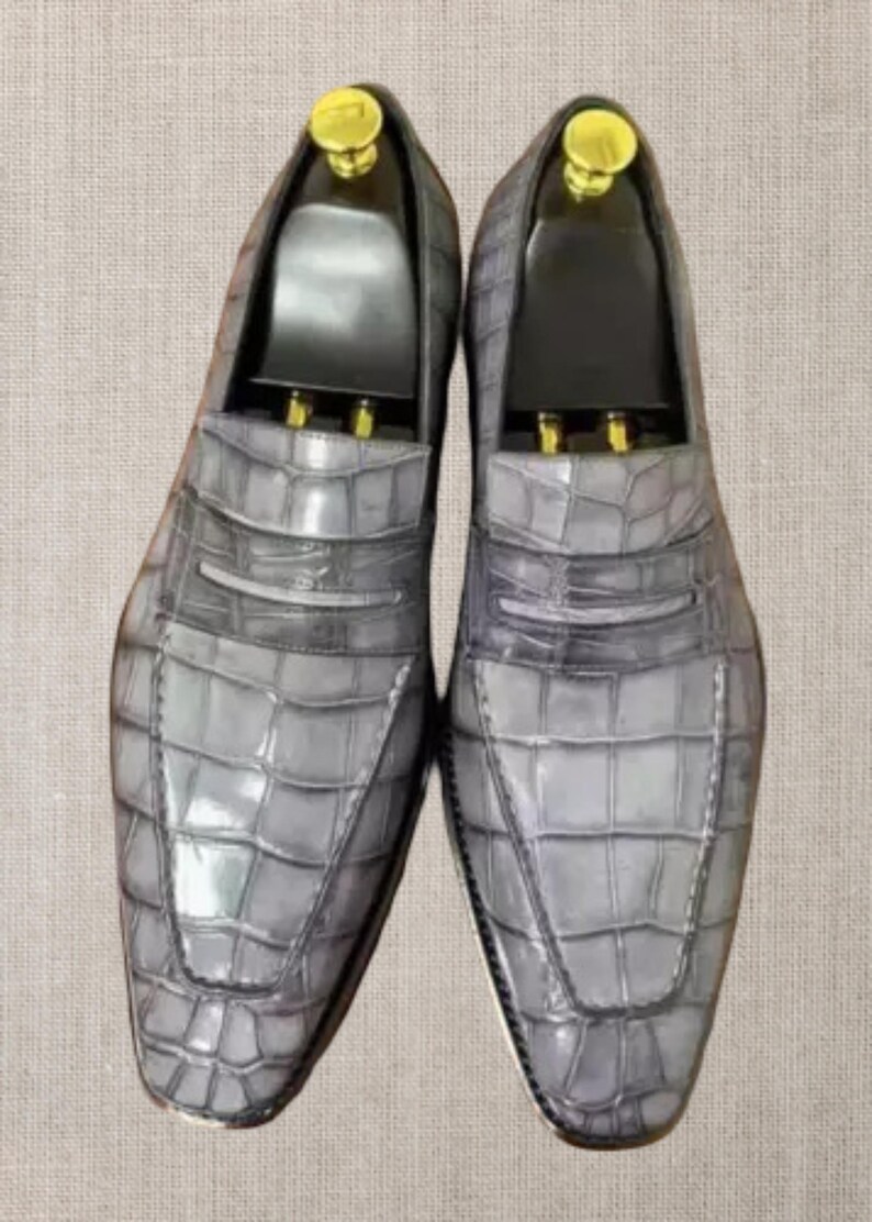 Op de afbeelding: Een paar grijze leren loafers met een krokodillenleertextuur. De schoenen hebben een klassiek ontwerp met een penny loafer-bandje en een ronde neus. Schoenbomen zijn ingevoegd.