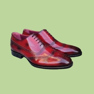 Pu&ograve; includere: Un paio di scarpe eleganti in pelle rossa con finitura lucida. Le scarpe presentano un classico design a coda di rondine con perforazioni decorative e lacci neri. Le suole sono nere, su sfondo verde chiaro.