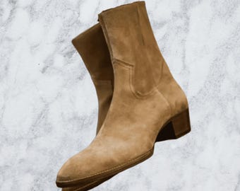 Puede incluir: Botines tobilleros de ante color beige con punta afilada y tacón cuadrado pequeño. Las botas tienen una cremallera en el lateral interior y un diseño cosido en la caña superior. Las botas son de color neutro.