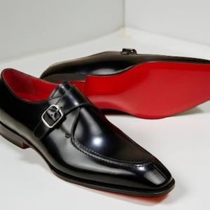 Peut inclure: Paire de chaussures noires en cuir à boucle, avec une finition brillante. Les chaussures présentent une boucle argentée et une semelle rouge. L'intérieur de la chaussure est également rouge. La seconde chaussure est floue en arrière-plan.