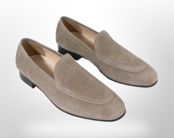 Puede incluir: Un par de mocasines de ante marrón claro con un diseño clásico sin cordones. Los zapatos tienen una puntera redondeada y un tacón bajo. Las suelas son negras. Los mocasines se muestran sobre un fondo blanco.