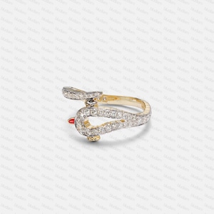 Op de afbeelding: Een gouden ring met een hoefijzervormig ontwerp, bezet met kleine, sprankelende diamanten. De ring heeft een rode accentkleur en een klein zwart detail, wat elegantie en stijl combineert.