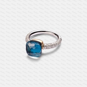 May include: A silver ring featuring a large, square-cut, blue gemstone with multiple facets. The ring's band is set with small, clear gemstones. The blue stone is set with a gold-coloured accent.