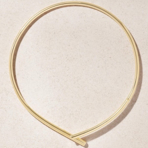 May include: A gold-tone choker necklace with a simple, modern design. The necklace forms a continuous loop, with a subtle overlap at the front. The metal has a polished finish, giving it a sleek and elegant appearance.