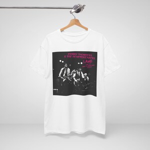 Peut inclure: Un t-shirt blanc à manches courtes avec une impression graphique en noir et blanc du groupe Johnny Thunders & The Heartbreakers. L'impression inclut le texte "Johnny Thunders & The Heartbreakers L.A.M.F. Live at the Lyceum Ballroom 1977" en rose.