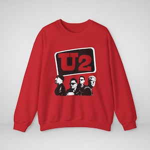 Puede incluir: Una sudadera roja de manga larga y cuello redondo con un estampado gráfico en el pecho. El diseño incluye el texto U2 en letras rojas negritas dentro de una forma rectangular negra con esquinas redondeadas, sobre una ilustración en blanco y negro de cuatro integrantes de la banda.