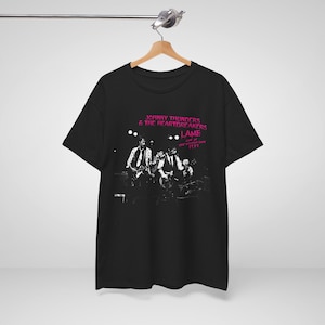 Puede incluir: Una camiseta negra con un estampado gráfico de la banda Johnny Thunders & The Heartbreakers. El estampado incluye el texto L.A.M.F. Live at the Village Gate 1977 en letras rosas sobre una fotografía en blanco y negro de la banda actuando.