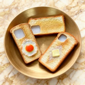 Puede incluir: Una colección de fundas para teléfono con diseño de tostadas. Una funda presenta un huevo frito, otra una porción de mantequilla y la tercera es lisa. Las fundas están dispuestas sobre un plato dorado.