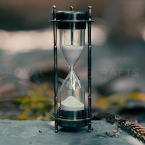 May include: An hourglass with a clear glass body and a silver-colored metal frame. The hourglass is filled with white sand. The background is blurred, with hints of green and brown foliage. The text "ROYAL CRAFTS" is visible.