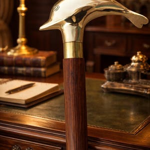 Puede incluir: Bastón con un mango de latón pulido en forma de delfín y un eje de madera oscura. El bastón tiene un collar de latón y un eje de madera oscura. El fondo muestra un escritorio con tapa de cuero verde, una lámpara de escritorio y libros.