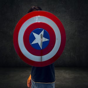 以下が含まれることがあります： 赤、白、青のキャプテン・アメリカの盾。盾の中央には銀色の星があり、青い円、白いリング、赤い外側のリングで囲まれています。盾は人の前に掲げられています。