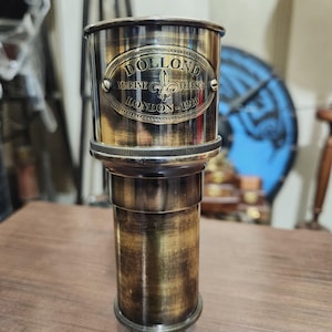 Puede incluir: Un telescopio de latón antiguo con el texto "DOLLOND MARINE TELESCO LONDON - 1920" grabado en la parte superior. El telescopio tiene forma cilíndrica con un diseño detallado y un acabado pulido. Es un instrumento náutico clásico.