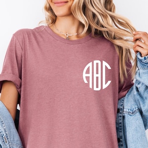 May include: A dusty rose-colored t-shirt with a white monogram of the letters "ABC" in a circular design. The shirt has rolled-up sleeves and is paired with a light blue denim jacket.