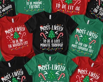 May include: A collection of Christmas-themed t-shirts in various colors, including black, red, and green. Each shirt features the phrase "MOST LIKELY" above a Christmas tree graphic and a humorous statement such as "to be a grinch".