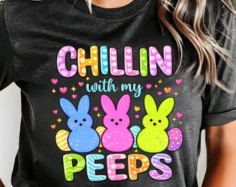 Peut inclure: T-shirt gris foncé avec l'inscription "CHILLIN with my PEEPS" en lettres colorées. Le motif présente trois lapins de Pâques de dessin animé bleus, roses et jaunes, avec des œufs de Pâques. Le t-shirt est de style décontracté et unisexe.