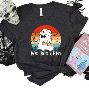 May include: Dark gray t-shirt featuring a cartoon ghost wearing a nurse's cap and a bandage, set against a sunset backdrop. The text "BOO BOO CREW" is below the graphic. Halloween-themed design.