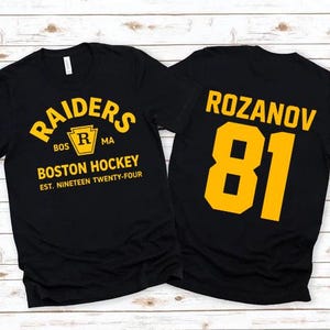 May include: Two black t-shirts with gold lettering. One shirt reads "RAIDERS BOSTON HOCKEY EST. NINETEEN TWENTY-FOUR" with a logo. The other shirt displays "ROZANOV 81".