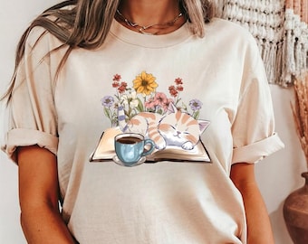Peut inclure: T-shirt beige avec un motif d'un chat tabby orange endormi sur un livre ouvert, entouré de fleurs colorées. Une tasse bleue avec du café est posée à côté du chat. Le t-shirt a des manches retroussées.