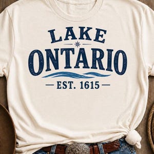 May include: A cream-colored short-sleeve t-shirt featuring dark blue text that reads LAKE ONTARIO with a small star icon, wavy lines, and the text EST. 1615 centered on the chest. The graphic has a distressed, textured appearance.