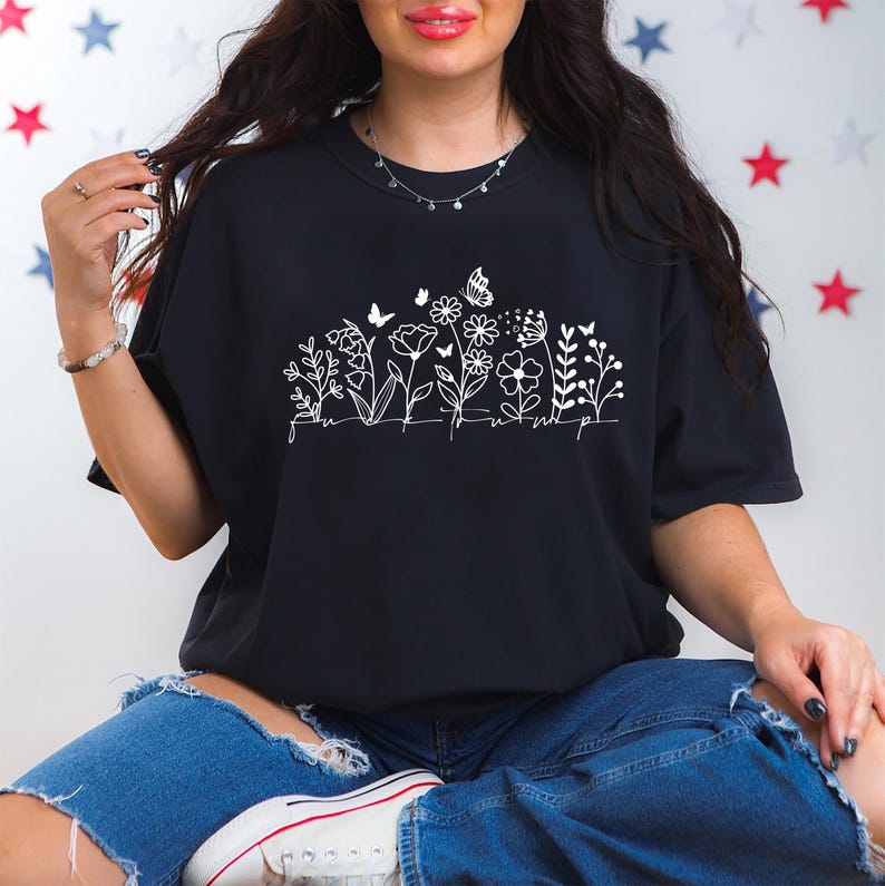 May include: Black t-shirt with a white floral design featuring butterflies and the word "flourish". The shirt is a crew neck and is paired with blue jeans. The overall theme is nature and botanical.