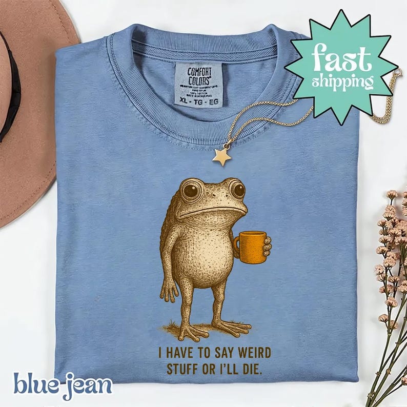 A blue t-shirt with a cartoon frog holding a coffee cup, accompanied by the text "I HAVE TO SAY WEIRD STUFF OR ILL DIE." The t-shirt is displayed on a white background, with a brown hat and some flowers in the background.