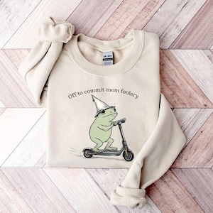 May include: Beige sweatshirt with the text "Off to commit mom foolery." The graphic shows a green frog wearing a wizard hat and riding a scooter. The sweatshirt is folded on a wooden surface.