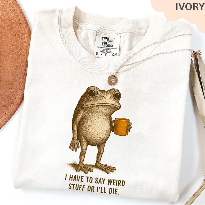 A white t-shirt with a printed image of a frog holding a cup, accompanied by the text "I HAVE TO SAY WEIRD Stuff or ILL DIE."