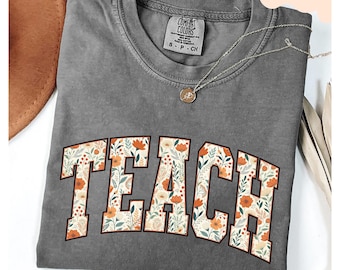 Peut inclure: Un t-shirt gris Comfort Colors avec le mot "TEACH" dans un motif floral. Les lettres sont remplies de fleurs orange, blanches et vertes. Un collier argenté avec un pendentif est drapé sur le t-shirt.