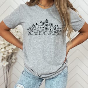 May include: A heather gray t-shirt featuring a black line drawing of flowers, butterflies, and the word "unwind" in a script font. The shirt has a crew neck and short sleeves.