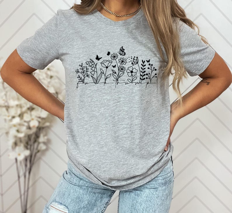 May include: A heather gray t-shirt featuring a black line drawing of flowers, butterflies, and the word "unwind" in a script font. The shirt has a crew neck and short sleeves.