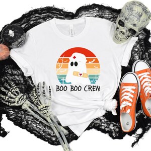 May include: White t-shirt with a graphic of a ghost wearing a nurse's cap and a bandage, set against a sunset design. The text "BOO BOO CREW" is below the graphic. Orange sneakers and Halloween decorations complete the scene.