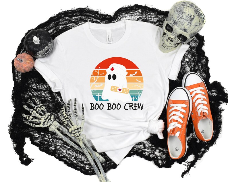 May include: White t-shirt with a graphic of a ghost wearing a nurse's cap and a bandage, set against a sunset design. The text "BOO BOO CREW" is below the graphic. Orange sneakers and Halloween decorations complete the scene.