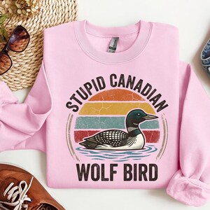 Może przedstawiać: R&oacute;żowa bluza z nadrukiem nurka na wodzie, na tle retro zachodu słońca. Tekst brzmi "STUPID CANADIAN WOLF BIRD". Bluza ma długie rękawy.