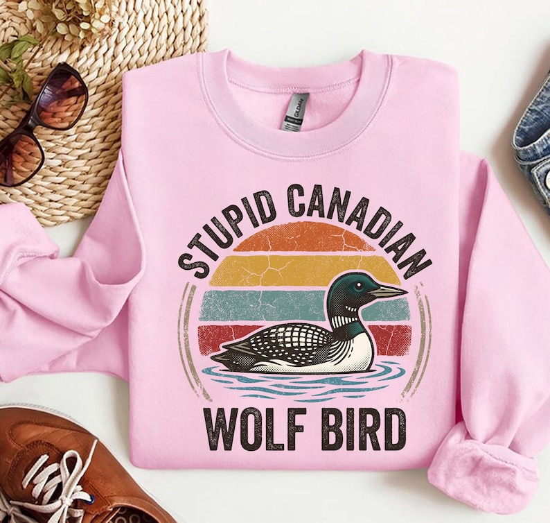 Może przedstawiać: R&oacute;żowa bluza z nadrukiem nurka na wodzie, na tle retro zachodu słońca. Tekst brzmi "STUPID CANADIAN WOLF BIRD". Bluza ma długie rękawy.