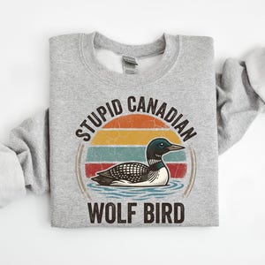 Może przedstawiać: Jasnoszara bluza z nadrukiem nurka na wodzie, na tle retro zachodu słońca. Tekst brzmi "STUPID CANADIAN WOLF BIRD" w okrągłym układzie. Bluza wykonana jest z miękkiego materiału.