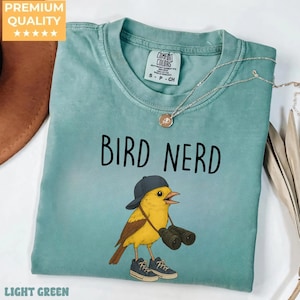 May include: Light green t-shirt with the words "BIRD NERD" above a cartoon bird wearing a cap, sneakers, and holding binoculars. The shirt has a crew neck and a small tag. A silver necklace with a pendant is also visible.