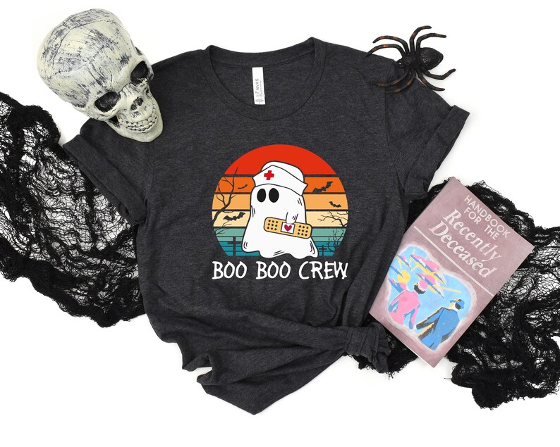 May include: Dark gray t-shirt featuring a cartoon ghost wearing a nurse's cap and a bandage, set against a sunset backdrop. The text "BOO BOO CREW" is below the graphic. Halloween-themed design.