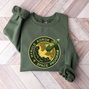 May include: Olive green sweatshirt featuring a circular graphic with the text "HYRULE KOROK SPACE PROGRAM." The graphic depicts a rocket ship launching from a yellow and green planet. The sweatshirt has long sleeves and a crew neck.