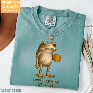 A light blue t-shirt with a cartoon frog holding a coffee cup and the text "I HAVE TO SAY WEIRD STUFF OR I'LL DIE." printed on it.