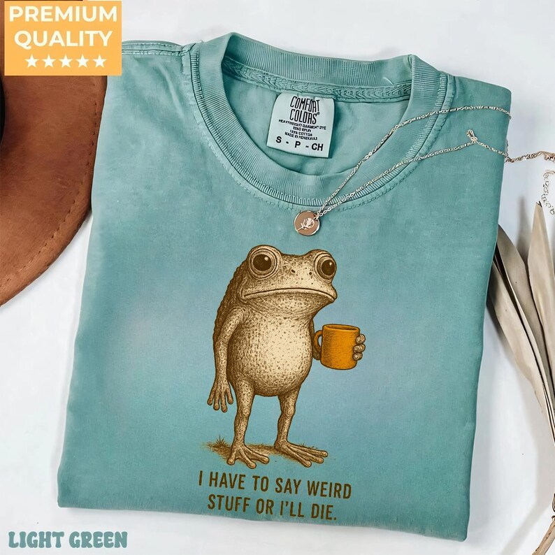 A light blue t-shirt with a cartoon frog holding a coffee cup and the text "I HAVE TO SAY WEIRD STUFF OR I'LL DIE." printed on it.