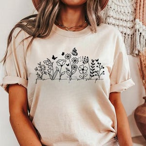 May include: A beige t-shirt featuring a black floral design with butterflies. The graphic includes various flowers and the word "sunshine" in a handwritten font. The shirt has rolled-up sleeves.