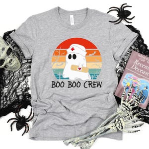 May include: A heather gray t-shirt featuring a cartoon ghost wearing a nurse's cap and holding a bandage, set against a retro sunset design. The text "BOO BOO CREW" is printed below the graphic. Halloween-themed apparel.