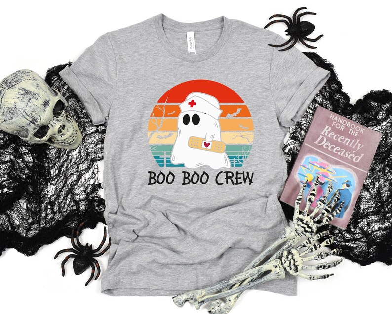 May include: A heather gray t-shirt featuring a cartoon ghost wearing a nurse's cap and holding a bandage, set against a retro sunset design. The text "BOO BOO CREW" is printed below the graphic. Halloween-themed apparel.
