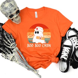 May include: Orange t-shirt with a graphic of a ghost wearing a nurse's cap and a bandage, set against a sunset design. The text "BOO BOO CREW" is below the graphic. Black and white sneakers are next to the shirt.